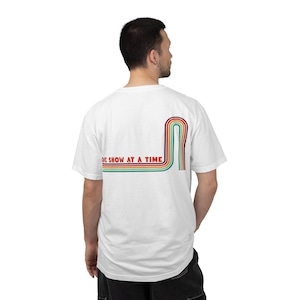 May include: White t-shirt with a retro rainbow design and the text "ONE SHOW AT A TIME" in red. The design features a curved, striped pattern in red, orange, yellow, green, and blue. The shirt is a classic crew neck style.