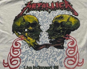 Puede incluir: Una camiseta blanca con un estampado gráfico de dos calaveras de color amarillo verdoso enfrentadas. Sobre las calaveras aparece el texto rojo METALLICA. Debajo hay dos formas rojas similares a serpientes y el texto negro Live in Concert 92.
