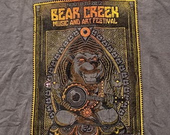 Puede incluir: Una camiseta gris carbón con un estampado gráfico del Bear Creek Music and Art Festival, 8-11 de nov. de 2012. El diseño muestra un oso central tocando el banjo, rodeado de intrincados patrones geométricos en dorado y negro.