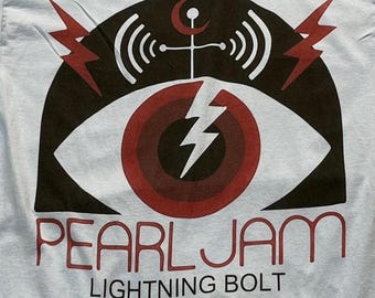 Puede incluir: Una camiseta azul claro con un estampado gráfico de un ojo grande con iris rojo y un rayo blanco en el centro. Sobre el ojo hay un arco negro con rayos rojos y un símbolo. El texto dice PEARL JAM LIGHTNING BOLT.