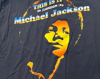 Puede incluir: Una camiseta negra con un estampado gráfico de Michael Jackson en tonos amarillos y naranjas. El texto dice THIS IS IT 50 | LONDON | 02 y Michael Jackson en una fuente con degradado azul y amarillo, sobre un contorno circular amarillo.