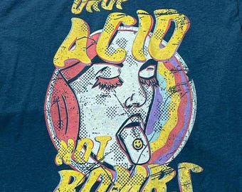 Puede incluir: Una camiseta azul cerceta oscuro con un estampado gráfico desgastado de un rostro con los ojos cerrados y la lengua fuera, sosteniendo una pequeña pastilla con una cara sonriente. El texto "DROP ACID NOT BOMBS" está impreso en amarillo con borde rojo.