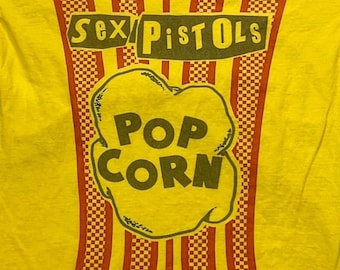 Puede incluir: Una camiseta de color amarillo brillante con un diseño gráfico de un cubo de palomitas con rayas verticales a cuadros rojos y blancos. El texto "SEX PISTOLS" aparece en la parte superior y las palabras "POP CORN" en el centro.