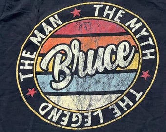 Puede incluir: Una camiseta negra con un gráfico circular sobre un fondo de atardecer desgastado en rojo, naranja, amarillo y azul. El texto "THE MAN THE MYTH THE LEGEND" rodea el perímetro, mientras que el nombre "Bruce" aparece en letra cursiva blanca.