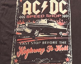 Puede incluir: Una camiseta negra con un estampado gráfico con el texto AC/DC SPEED SHOP y LAST STOP BEFORE THE HIGHWAY TO HELL. El diseño incluye un coche antiguo, llamas y el texto MAKING SERIOUS HORSEPOWER SINCE 1973 en un estilo desgastado.