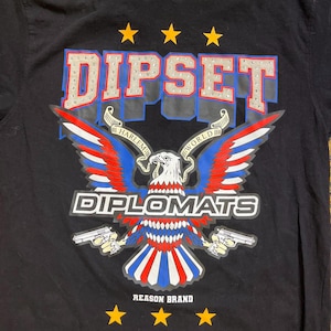 Puede incluir: Camiseta negra con un diseño gráfico. El diseño incluye la palabra "DIPSET" en plata y azul, sobre un águila con alas rojas, blancas y azules. La palabra "DIPLOMATS" está debajo del águila, y "REASON BRAND" en la parte inferior.