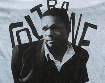 Puede incluir: Una camiseta de color claro con una impresión fotográfica en blanco y negro de una persona con una chaqueta de traje oscura y una camisa de cuello claro. El texto COLTRANE está impreso en letras mayúsculas negras, grandes y negritas.