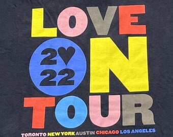 Puede incluir: Una camiseta negra con el texto colorido LOVE ON TOUR, el año 2022 y un símbolo de corazón dentro de un círculo azul. Debajo del gráfico principal, los nombres de las ciudades Toronto, New York, Austin, Chicago y Los Angeles están en letra pequeña.