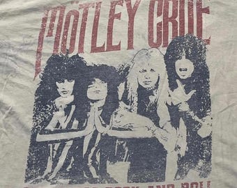 Puede incluir: Una camiseta de color claro con un gráfico oscuro desgastado de la banda Mötley Crüe. El estampado muestra a cuatro miembros de la banda con el pelo largo y el texto MÖTLEY CRÜE arriba, con la frase SEX DRUGS ROCK AND ROLL impresa debajo.