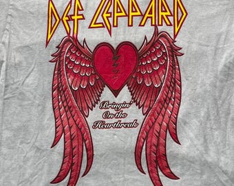 Puede incluir: Una camiseta gris claro con un estampado gráfico de un corazón rojo con una grieta en zigzag en el centro, flanqueado por dos grandes alas rojas con plumas. Sobre el corazón está el texto Def Leppard y debajo Bringin On the Heartbreak.