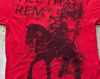 Puede incluir: Una camiseta roja de manga corta con un estampado gráfico oscuro. El diseño muestra una figura esquelética montando un caballo y sosteniendo un bastón alto, con el texto ALL THAT REMAINS impreso en una fuente negra desgastada sobre la ilustración.
