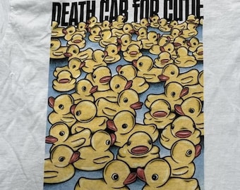 Puede incluir: Una camiseta blanca con un estampado gráfico de numerosos patitos de goma amarillos flotando sobre una superficie azul. Sobre los patos, el texto DEATH CAB FOR CUTIE está impreso en letras mayúsculas negras y negritas.