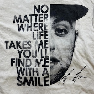 May include: A cream-coloured t-shirt with a black and white portrait of a man and the text "NO MATTER WHERE LIFE TAKES ME YOU'LL FIND ME WITH A SMILE." The shirt also includes a signature.
