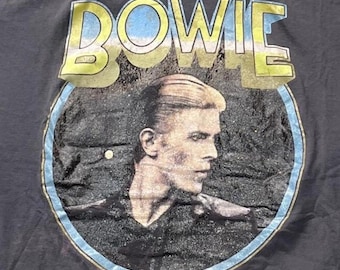 Puede incluir: Una camiseta gris oscuro con un gráfico circular de David Bowie de perfil. La palabra BOWIE está impresa en letras amarillas grandes y desgastadas sobre el círculo. El diseño incluye un fondo oscuro texturizado y un borde circular azul claro.