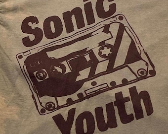 Puede incluir: Una camiseta de color verde oliva claro con un estampado gráfico en marrón oscuro de un casete. El texto Sonic está impreso sobre la ilustración del casete, y el texto Youth está impreso debajo en una fuente oscura y en negrita.