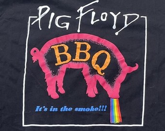 Puede incluir: Una camiseta negra con un gráfico de la silueta de un cerdo rosa con el texto naranja BBQ dentro. Sobre el cerdo está el texto blanco PIG FLOYD, y debajo el texto azul It's in the smoke!!! Una estela de arcoíris sale de la cola del cerdo.