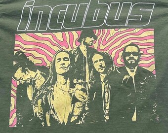 Puede incluir: Una camiseta verde oliva con un estampado gráfico de la banda Incubus. El diseño incluye la palabra "incubus" en letras blancas con borde sobre una imagen rectangular de cuatro miembros de la banda con un fondo ondulado rosa y beige.