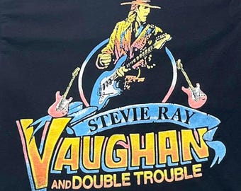 Puede incluir: Una camiseta negra con un gráfico de un guitarrista con sombrero tocando una guitarra eléctrica. El diseño incluye dos guitarras adicionales y una pancarta azul con el texto STEVIE RAY VAUGHAN AND DOUBLE TROUBLE en amarillo y naranja.