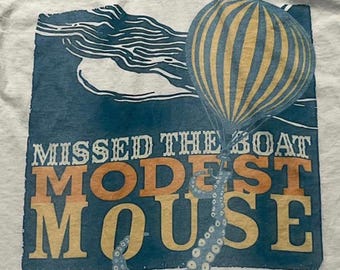 Puede incluir: Una camiseta gris claro con un estampado gráfico sobre fondo azul. El diseño muestra un globo aerostático amarillo y azul sujeto por un tentáculo de pulpo, con el texto MISSED THE BOAT MODEST MOUSE impreso en blanco y naranja.