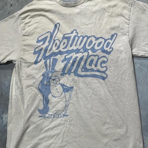 May include: A light beige t-shirt with the words "Fleetwood Mac" in light blue script above a cartoon penguin in a top hat, bow tie, and cane. The penguin is also light blue.