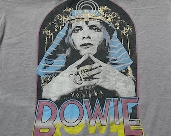 Puede incluir: Una camiseta gris claro con un estampado gráfico de una persona con un tocado y joyas de estilo egipcio. La persona tiene las manos juntas cerca de la cara. Debajo de la imagen, la palabra BOWIE está impresa en letras grandes y coloridas.