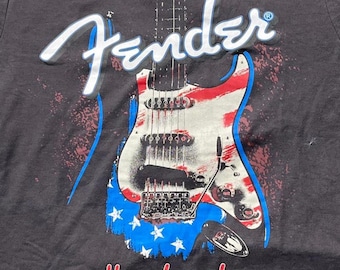 Puede incluir: Una camiseta negra con el logotipo de Fender en blanco en la parte superior y un gráfico de una guitarra eléctrica con un patrón de bandera estadounidense en rojo, blanco y azul. La palabra Handmade está impresa en rojo en la parte inferior.