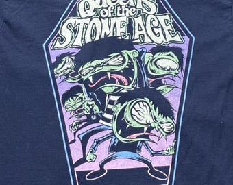 Puede incluir: Una camiseta azul oscuro con un estampado gráfico dentro de un contorno en forma de ataúd. El diseño muestra el texto Queens of the Stone Age en letras blancas sobre tres criaturas estilizadas de piel verde y dientes afilados.