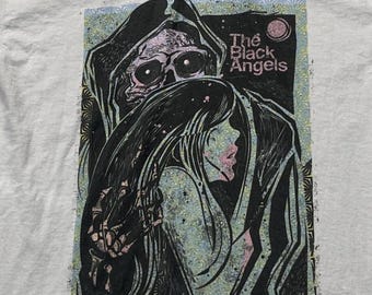 Puede incluir: Una camiseta gris claro con un estampado gráfico de una figura esquelética con capucha oscura abrazando a una persona de pelo largo. El texto The Black Angels está impreso en rosa sobre las figuras. El diseño usa negro, rosa y azul claro.