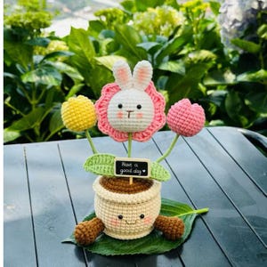 May include: A handmade crochet flower arrangement. It features a bunny face with pink petals, yellow and pink tulips, and a pot with a smiling face. A small chalkboard sign reads "Have a good day". The arrangement is set against green foliage.