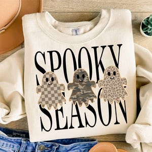 May include: A cream-colored crewneck sweatshirt featuring the black text SPOOKY SEASON. Below the text are three ghost-shaped graphics: one with a tan checkerboard pattern, one with a green and brown camouflage print, and one with a tan leopard print.