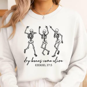 Puede incluir: Una sudadera blanca de cuello redondo con un gráfico negro de tres esqueletos bailando. Debajo de los esqueletos, el texto dice "dry bones come alive" en letra cursiva, seguido del texto impreso "EZEKIEL 37:5" en una fuente sencilla.