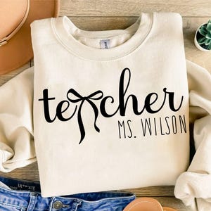 May include: Cream-colored sweatshirt with the word "teacher" in a black script font, featuring a bow design. Below the word "teacher" is the name "Ms. Wilson" in a smaller, block font. The sweatshirt is folded on a wooden surface.