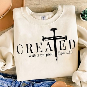 May include: A cream-colored crewneck sweatshirt featuring black text that reads CREATED with a purpose Eph 2:10, where the letter T is replaced by a graphic of a large nail shaped like a cross. The garment has long sleeves and a ribbed neckline.