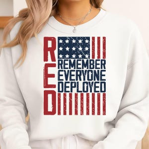 Può includere: Felpa bianca con un design patriottico. Il design presenta la parola "RED" in rosso, con una bandiera americana blu e bianca e le parole "REMEMBER EVERYONE DEPLOYED" in blu.