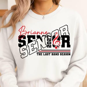 May include: A white crewneck sweatshirt featuring a graphic design with the red script text Brianne, the black block text SENIOR, the vertical text 2027, a red treble clef, and the black text THE LAST BAND SEASON with red and black lines.