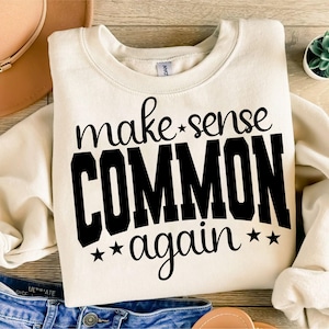 May include: Cream-colored sweatshirt with the phrase "make sense COMMON again" in black lettering. The word "COMMON" is in a bold, block font. The sweatshirt is folded on a wooden surface, with a hat and jeans visible.