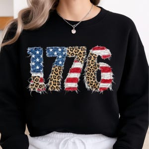 May include: Black sweatshirt with the year "1776" printed on the front. The numbers are designed with American flag and leopard print patterns. A casual top, the sweatshirt is a comfortable garment for everyday wear.
