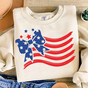 May include: A cream-colored crewneck sweatshirt featuring a graphic of a large blue bow patterned with white stars, accompanied by five red horizontal brushstroke stripes and several small red and blue stars scattered above the bow design.
