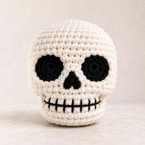 Puede incluir: Una calavera tejida a ganchillo a mano con hilo color blanco roto. La calavera presenta dos grandes cuencas oculares circulares de hilo negro, una pequeña cavidad nasal triangular negra y una línea negra cosida que representa la boca.