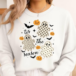 Könnte beinhalten: Ein weißes Sweatshirt mit Rundhalsausschnitt und einem Grafikdruck mit dem Text 'tis the season' in schwarzer Schreibschrift. Das Design zeigt drei Geister mit verschiedenen Mustern: kariert, Leopardenmuster und gepunktet, umgeben von orangefarbenen Kürbissen und schwarzen Fledermäusen.