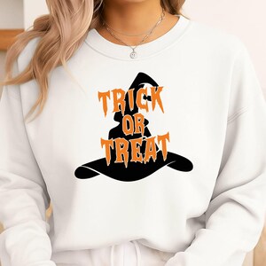 May include: A white crewneck sweatshirt featuring a black graphic of a witch hat. The orange text "TRICK OR TREAT" is printed across the center of the hat in a jagged, stylized font. The sweatshirt has long sleeves and a ribbed neckline.