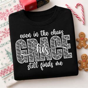 May include: A black crewneck sweatshirt featuring white text that reads "even in the chaos his grace still finds me." The word "GRACE" is written in large, bold, block letters filled with a detailed white floral pattern on a black background.