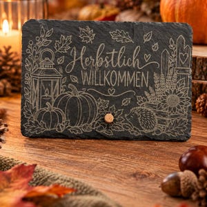 May include: A rectangular black slate plaque featuring an engraved design of pumpkins, a lantern, a hedgehog, and autumn leaves. The text "Herbstlich WILLKOMMEN" is centred on the surface. A small wooden knob is attached to the lower centre.