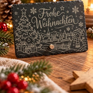 May include: A rectangular black slate plaque featuring the engraved text Frohe Weihnachten, a decorated Christmas tree with gifts, a winter village scene with a church, and a snowman. A small wooden peg is centred near the bottom edge.