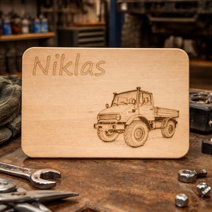 May include: A light brown wooden chopping board engraved with the name "Niklas" and a truck design. The rectangular board has rounded corners. The background shows a workshop setting with tools and a vehicle.