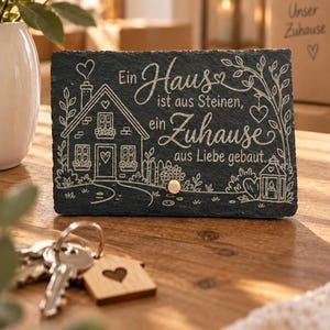 May include: A rectangular black slate plaque featuring an engraved illustration of a house, a tree, and a lantern. The text reads: Ein Haus ist aus Steinen, ein Zuhause aus Liebe gebaut. A small circular gold-coloured metal knob is attached to the front.