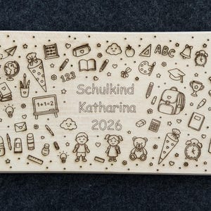 May include: A light brown rectangular wooden plaque featuring engraved school-related illustrations such as a backpack, pencils, and a mortarboard. The text "Schulkind Katharina 2026" is also engraved on the plaque. The plaque is light brown.