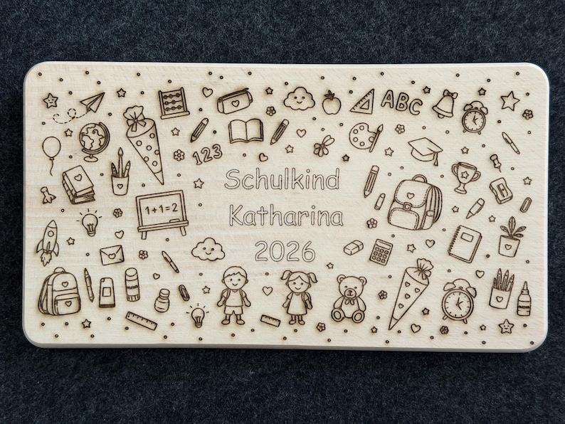 May include: A light brown rectangular wooden plaque featuring engraved school-related illustrations such as a backpack, pencils, and a mortarboard. The text "Schulkind Katharina 2026" is also engraved on the plaque. The plaque is light brown.