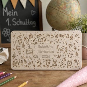 May include: A wooden plaque engraved with school-related icons, such as a backpack, pencils, and a globe. The plaque is personalised with the name "Katharina" and the year "2026". A chalkboard with text and a globe are in the background.