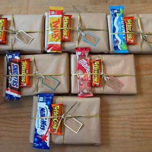 May include: Several gifts wrapped in brown paper, tied with twine, and adorned with small tags. Each package is decorated with a candy bar, including Snickers, Twix, Bounty, and Milky Way. The gifts are arranged on a wooden surface.